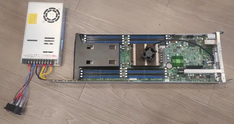 A top-view image displaying a blade server motherboard connected via a custom adapter to a standalone enclosed power supply unit (PSU). The setup shows connections and cables organized between the PSU terminals and the motherboard.