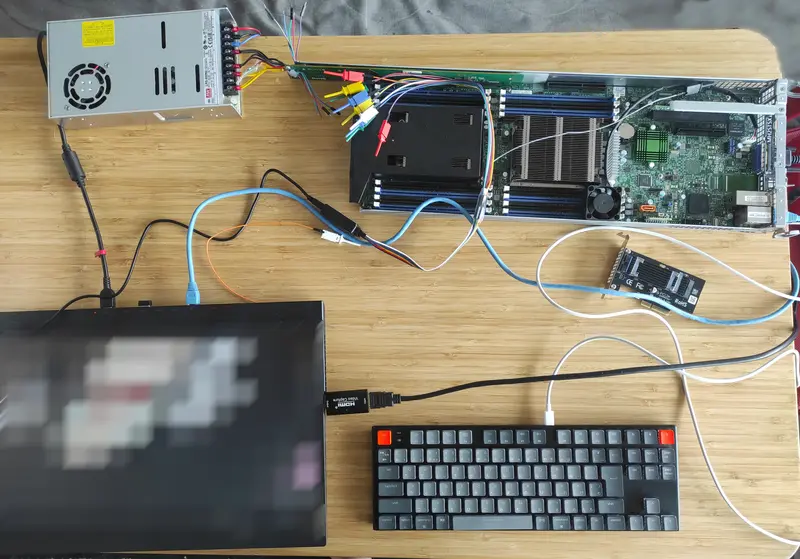 A complete test setup on a wooden tabletop showing a blade server motherboard connected via custom wiring to a standalone power supply unit. Additional cables connect peripherals including a keyboard, external SSD, Ethernet cables, and a laptop (display blurred).