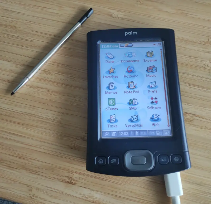 A Palm TX personal digital assistant (PDA) with stylus beside it, display is on and main application menu and connected to a USB cable.