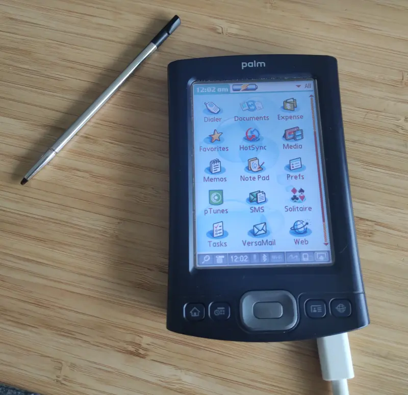 A Palm TX personal digital assistant (PDA) with stylus beside it, display is on and main application menu and connected to a USB cable.