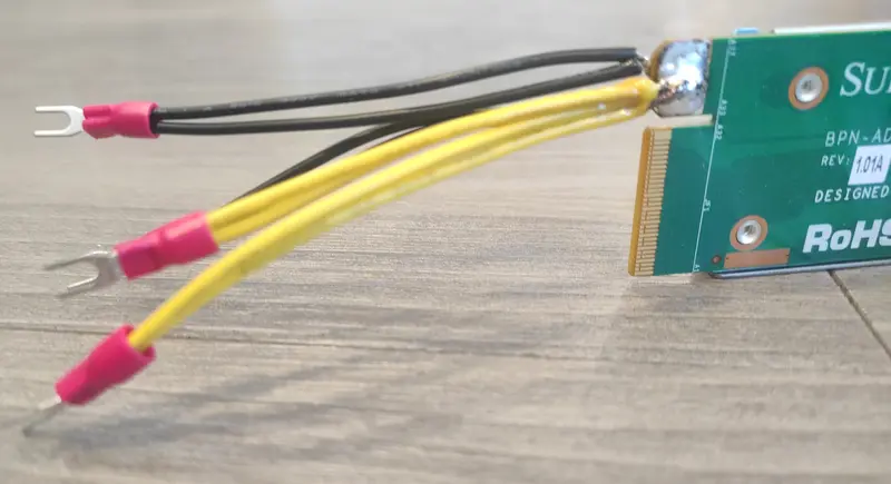 A close-up photo showing a modified backplane PCB with black and yellow wires soldered onto the edge connector pads. The wires end with red insulated fork terminals, indicating custom wiring for power connector.